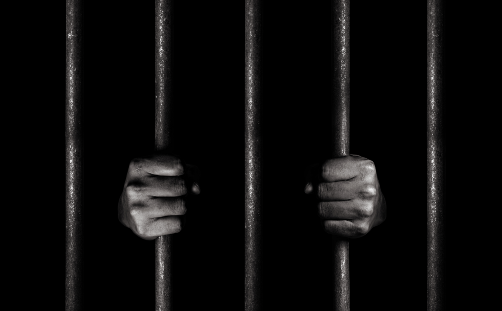 A man with dirty hands tightly gripping the bars of a jail cell.